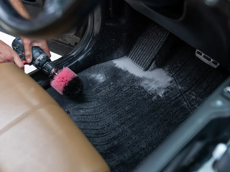 Drill brush working cleaner into a black floor mat next to a tan leather seat